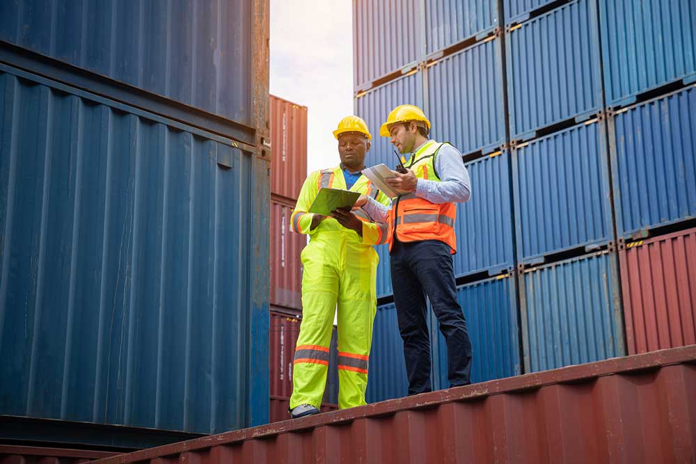 Two men in safety vests stand on shipping containers, overseeing global trade operations in a busy port area.