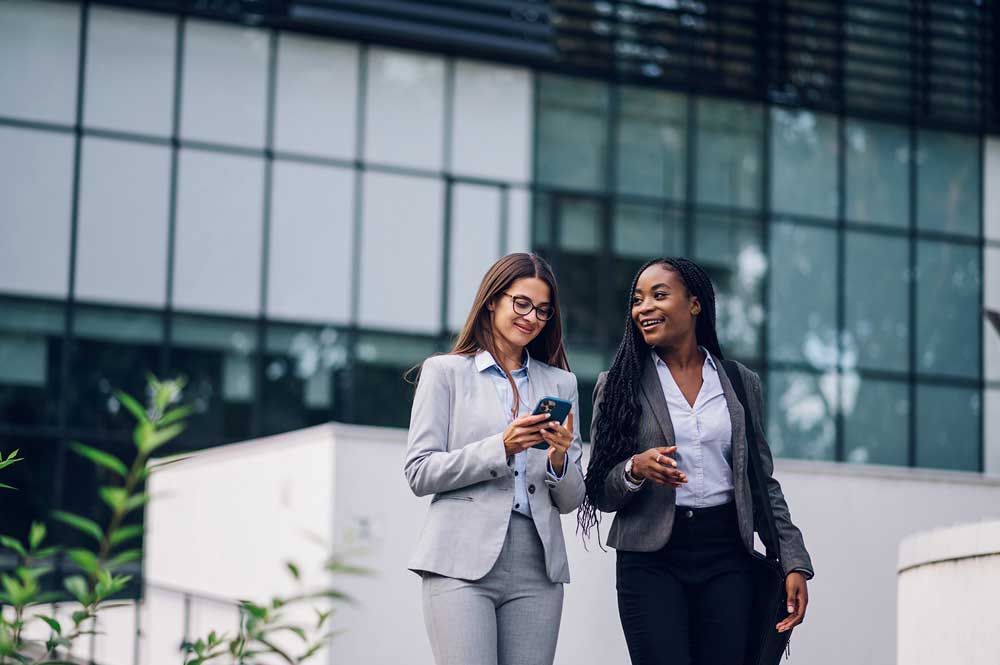 Two women strolling side by side in front of a corporate building.